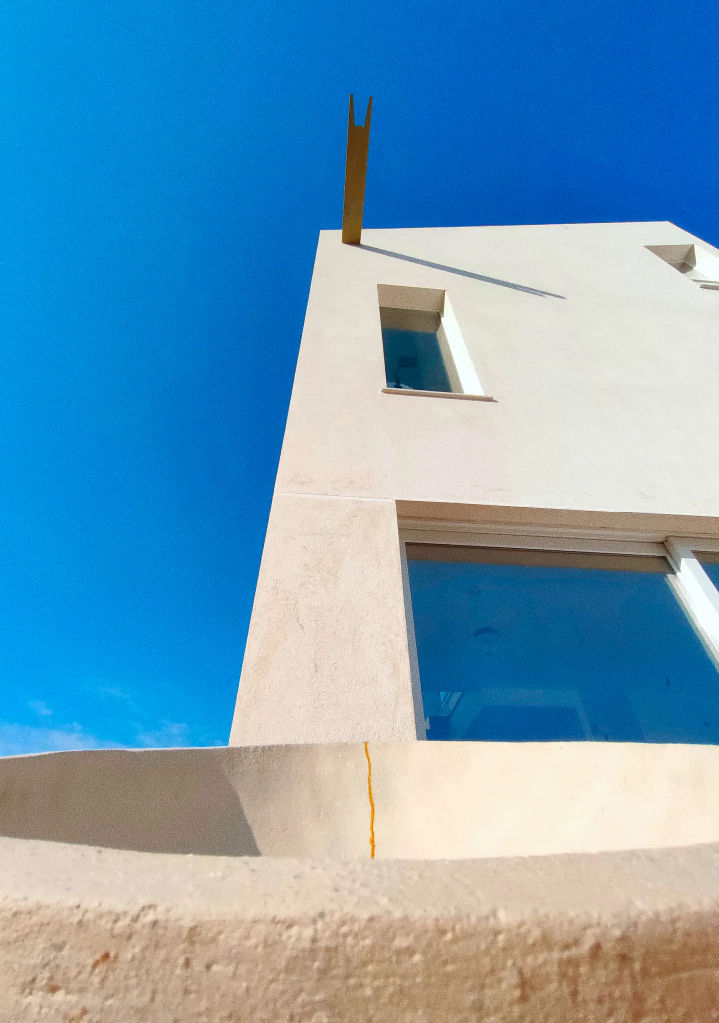 Casa de arquitectura minimalista con fachada en color blanco que destaca contra un cielo azul claro. Una estrecha ventana y una viga sobresalen de la fachada, creando una sensación de elegancia y amplitud y conectan focalmente con un recipiente blanco y dorado.