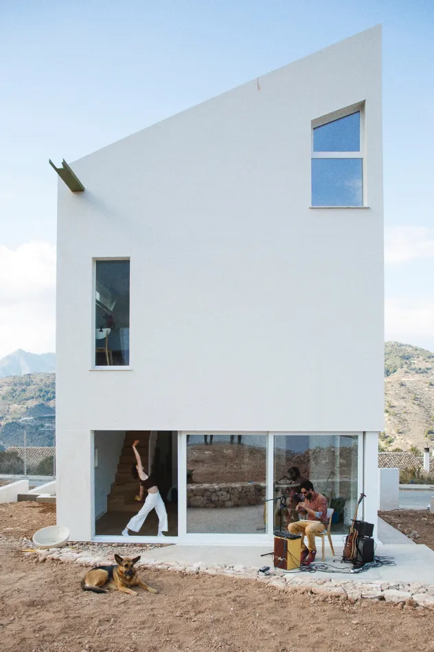 La casa blanca, moderna y minimalista con grandes ventanales, se enmarca en un paisaje rocoso. Una persona baila en el interior, junto al marco de la puerta, mientras otra toca música en el exterior. Un perro descansa cerca de allí.