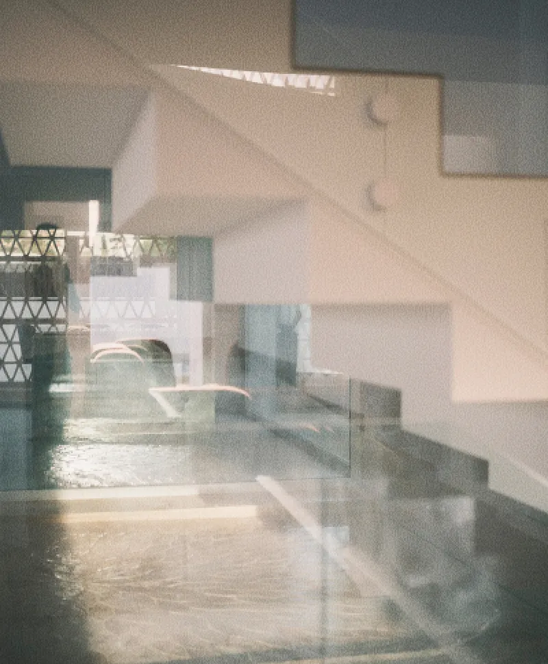 Fotografía evocadora de unas escaleras blancas y, tras ellas, un interior arquitectónico minimalista. Sobre el cristal que precede a la escena se refleja una celosía con patrones geométricos y agua.