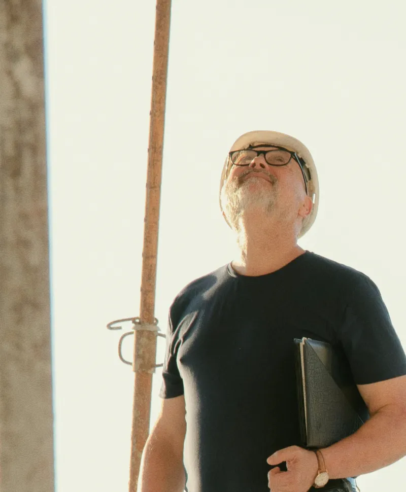 Serafín Galiano con casco de seguridad blanco mirando hacia arriba con expresión de satisfacción en una obra en construcción mientras sostiene una carpeta.