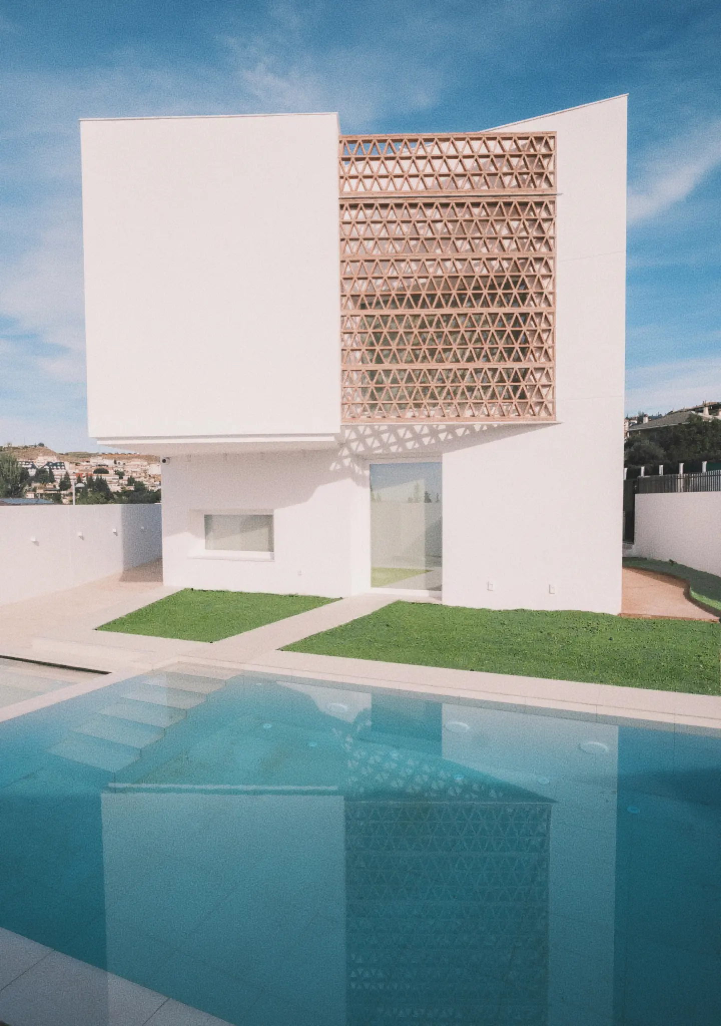 Casa moderna de color blanco con celosía geométrica, que se refleja en una piscina de aguas cristalinas. El césped verde rodea la piscina, bajo un cielo luminoso y con alguna nube.