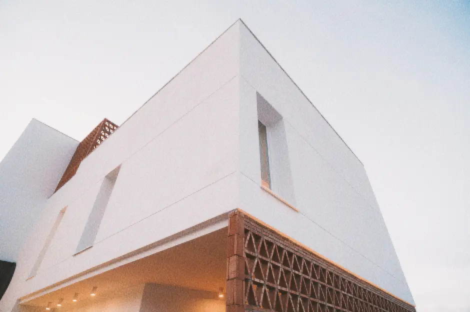 Exterior de la vivienda, de color blanco con líneas geométricas, caracterizada por un diseño en celosía y grandes ventanas verticales. El cielo está despejado, lo que transmite un ambiente minimalista y sereno.