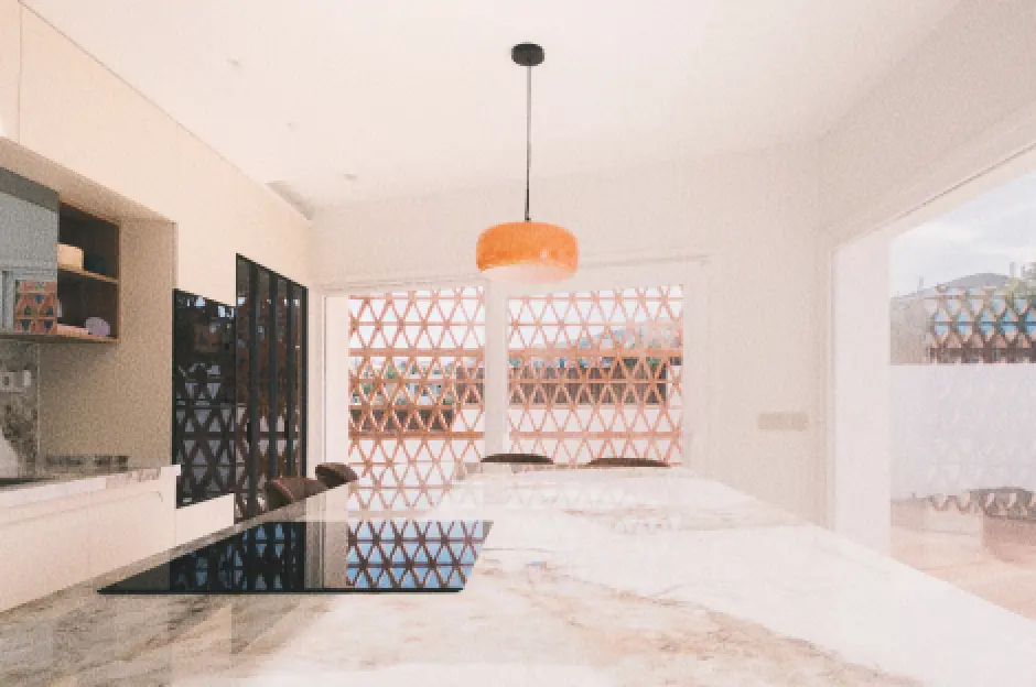 Cocina moderna con una elegante isla de mármol, una placa de inducción negra y una lámpara colgante naranja. Las ventanas grandes y con celosía dejan pasar abundante luz natural.