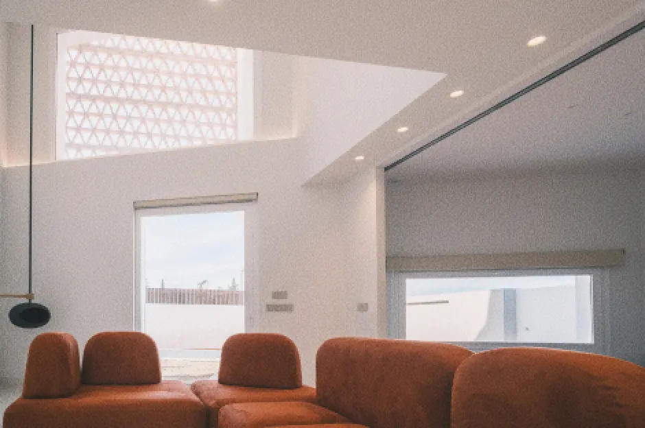 Un salón moderno y minimalista con techos altos, en el que destaca un sofá modular de color naranja. Los grandes ventanales y la claraboya que conecta con la celosía exterior aportan abundante luz natural.