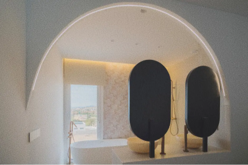 Cuarto de baño moderno con dos espejos ovalados negros, una bañera exenta con grifería dorada y un gran ventanal. La iluminación tenue y la abertura en arco crean un ambiente sereno.