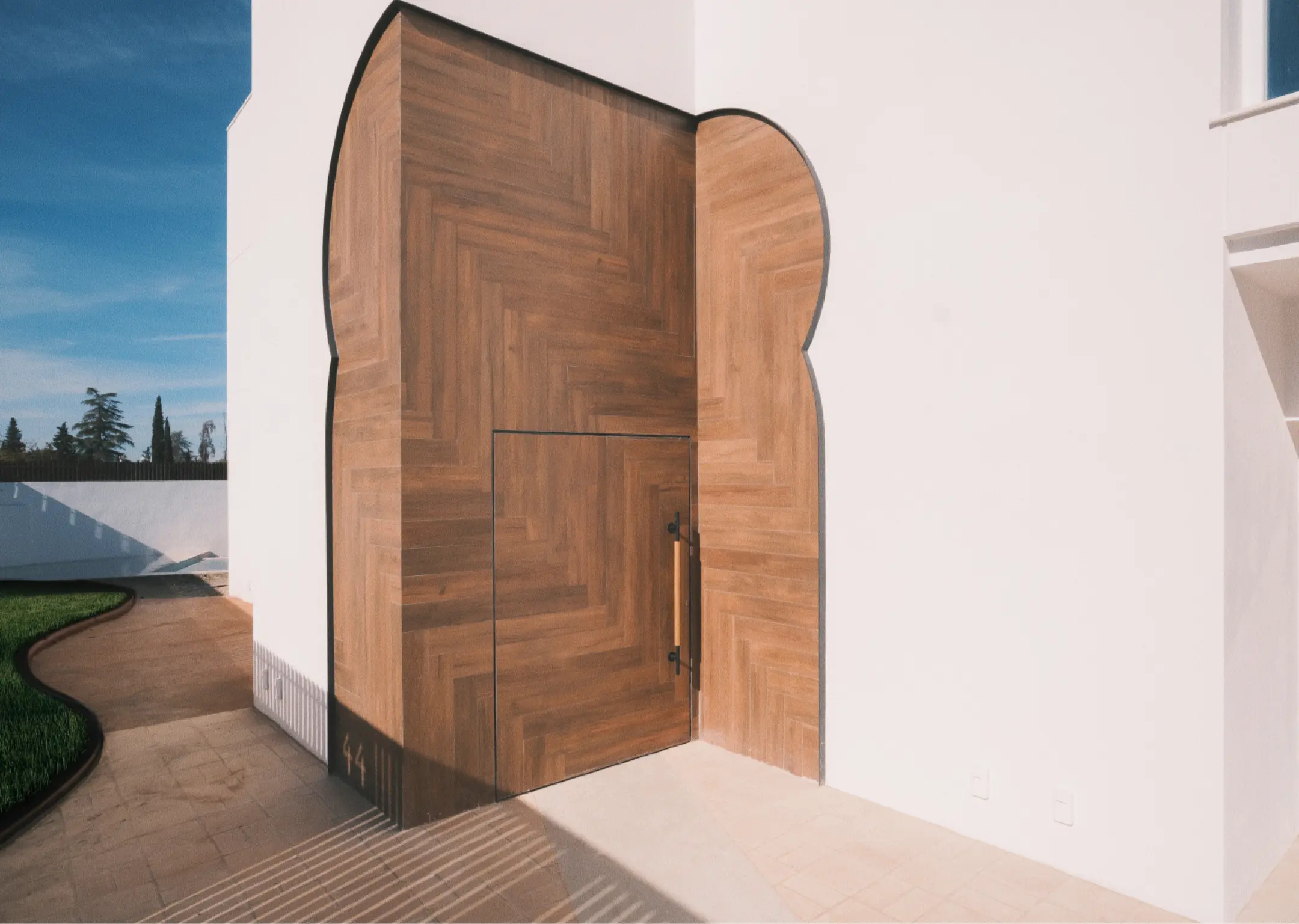 Una gran puerta de madera de formas abstractas contrasta con una pared blanca bajo un cielo azul claro, transmitiendo una sensación de diseño arquitectónico moderno con claras referencias a la arquitectura andalusí.