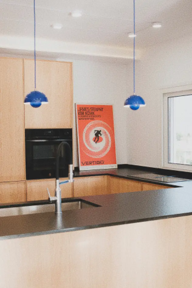 Cocina moderna con armarios de madera, encimeras negras y lámparas colgantes azules. Un póster de gran formato de la película Vértigo (A. Hitchcock) está apoyado contra la pared, cerca de una ventana.