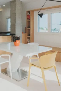 Comedor con una elegante mesa blanca con pie metalizado, una maceta naranja como centro de mesa y sillas amarillo pastel y blancas. Al fondo hay una ventana rodeada por una estantería y un banco de carpintería. La gran ventana y la decoración minimalista crean una sensación de amplitud.