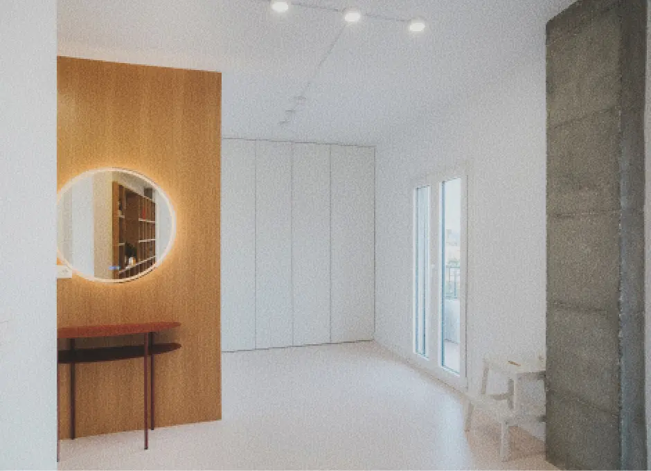 Habitación minimalista de acceso al vestidor y baño, con un panel de madera del que cuelga un espejo redondo retroiluminado y una mesita en madera más oscura. Las paredes blancas, la columna de hormigón visto, las lámparas de techo y la puerta del balcón aportan luminosidad.