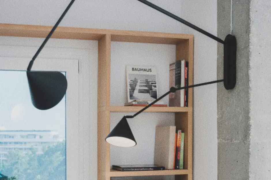 Detalle del salón-comedor con una estantería de madera, libros, una revista «Bauhaus», un gran ventanal y una elegante lámpara de pared negra que se bifurca en dos focos y que se extiende sobre la escena.