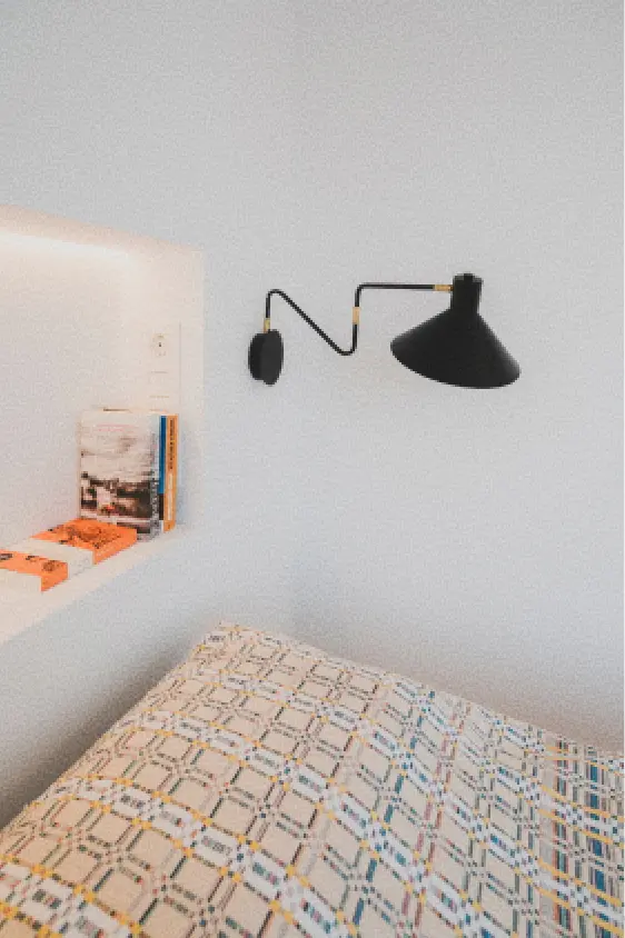 Un rincón acogedor en el dormitorio con un aplique negro, ropa de cama estampada y una hornacina con libros que funciona como cabecero. El ambiente es sereno y minimalista.