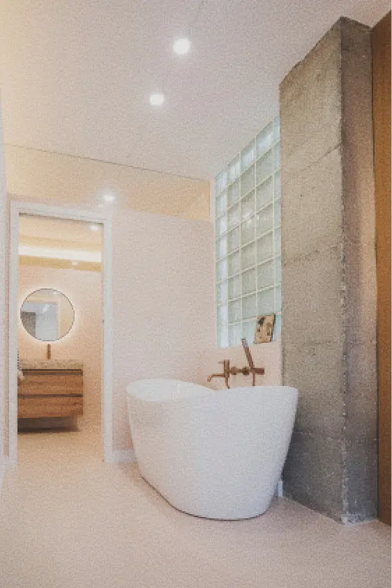 Un cuarto de baño moderno con una bañera exenta blanca con grifería en oro cepillado, un tabique decorativo de hormigón, una ventana de bloques de vidrio y un tocador minimalista con un espejo redondo, que crean un ambiente relajado.