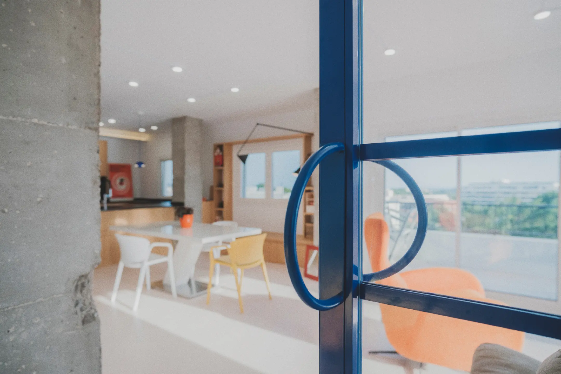Salón-comedor con cocina americana visto a través de una puerta acristalada con marco azul, en el que destacan un juego de comedor blanco y amarillo, sillones naranjas y grandes ventanales con vistas soleadas.