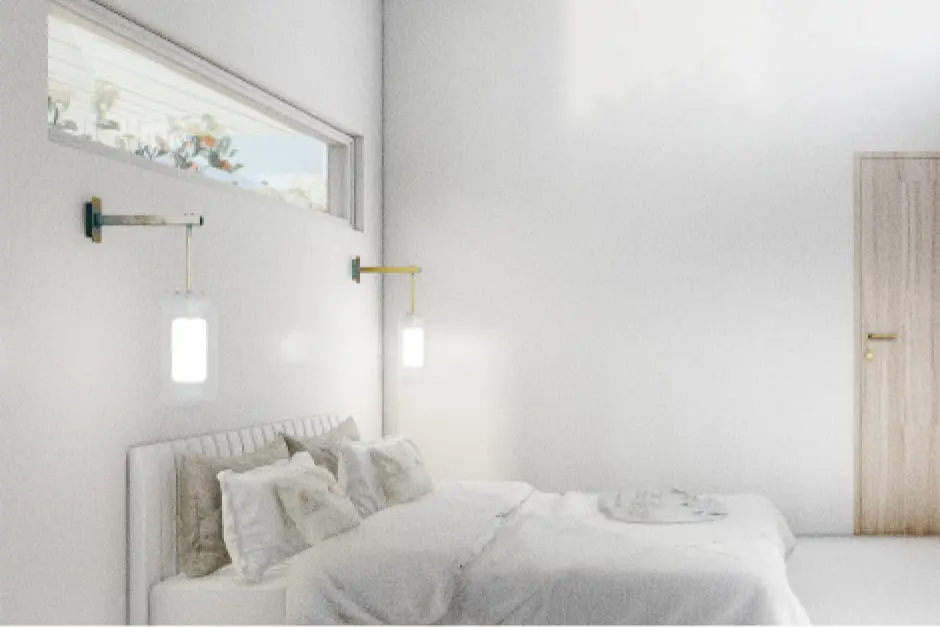 Dormitorio amplio con paredes blancas, una suave luz natural, una cama con cojines y lámparas de pared colgantes a los laterales. Una pequeña ventana situada sobre la cama le da un toque de serenidad y aporta iluminación natural.
