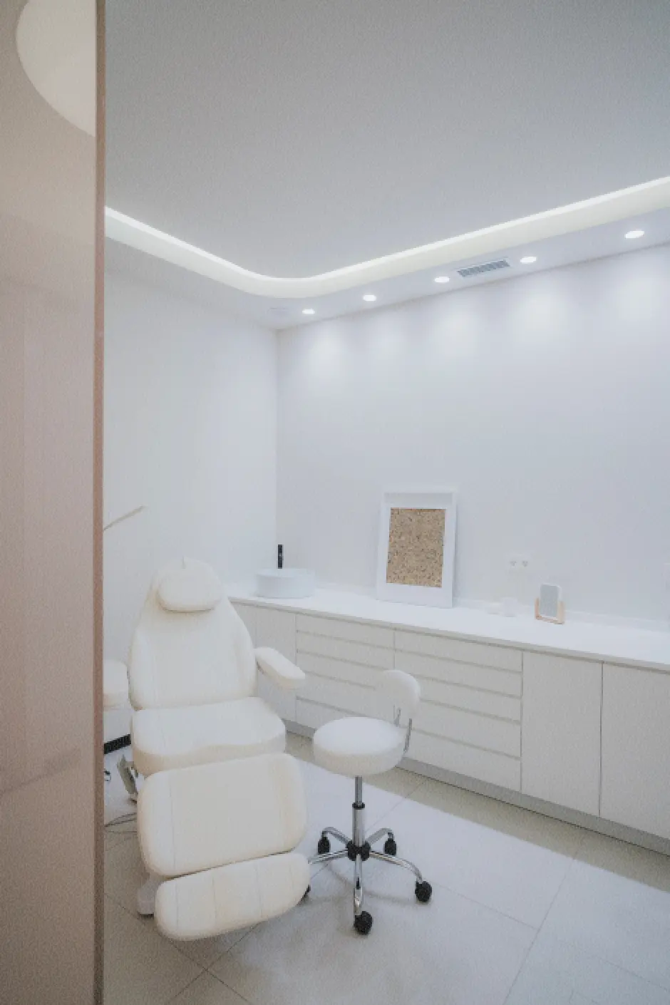 Consulta médica luminosa y moderna, con paredes blancas, armarios de líneas elegantes, un sillón de exploración y un taburete pequeño. Ambiente limpio, minimalista y tranquilo.