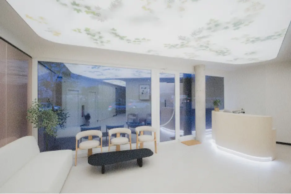 Una sala de espera moderna con techo traslúcido retroiluminado a través del cual se observan ramas y hojas, un mostrador de recepción curvo, sofá y sollas en blanco y madera, y grandes ventanales de fachada. Transmite tranquilidad y pulcritud.