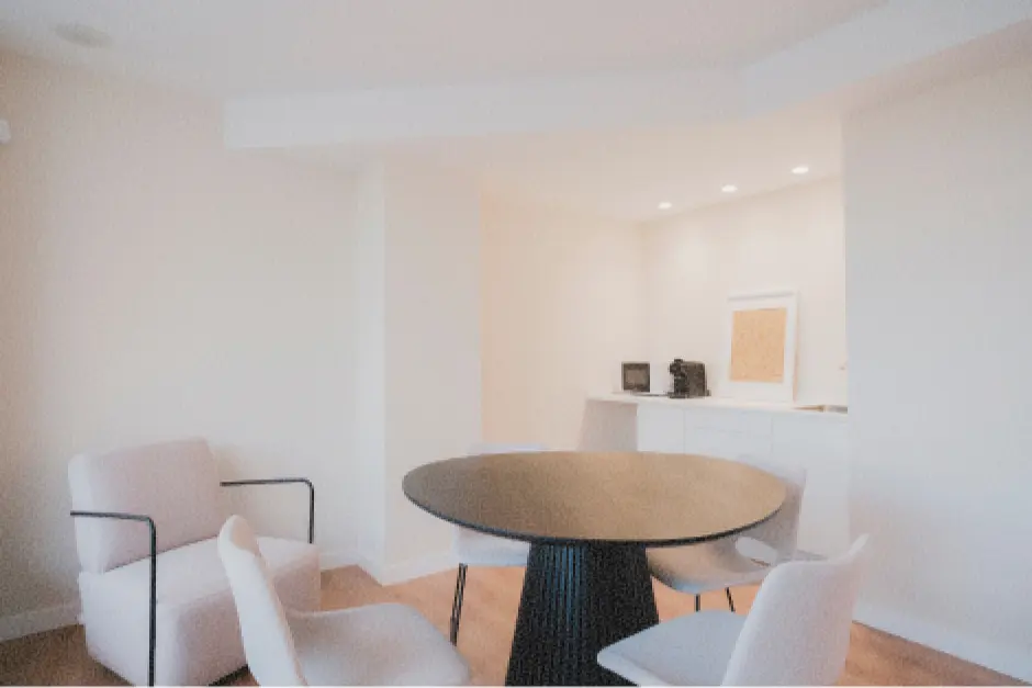 Cocina minimalista con una mesa redonda negra, cuatro sillas blancas y un rincón acogedor con una iluminación suave donde se encuentra la cafetera y el fregadero sobre muebles bajos. Ambiente moderno y tranquilo.