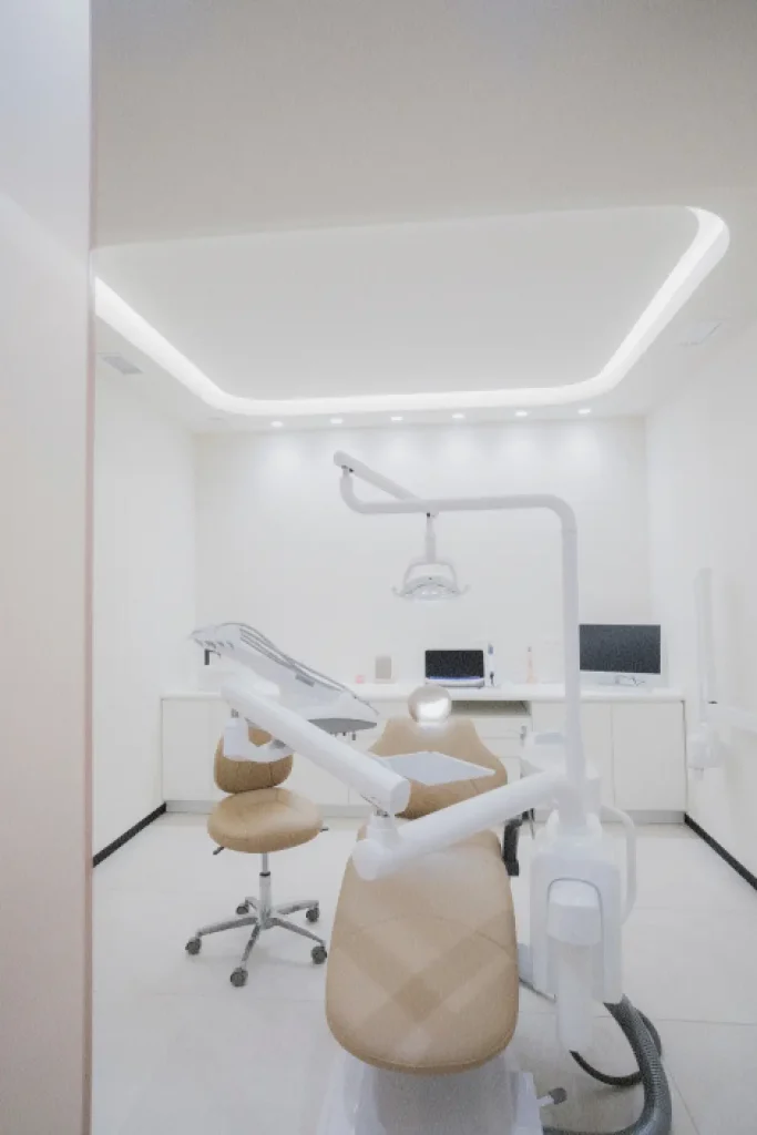 Consulta dental con un sillón dental de color beige, una lámpara de techo y el equipo necesario. Diseño luminoso y limpio, con una estética minimalista y varios monitores sobre el mostrador.